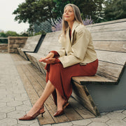 A woman sits on a wooden outdoor bench wearing a beige blazer, rust-red skirt, and brown pointed-toe heels.