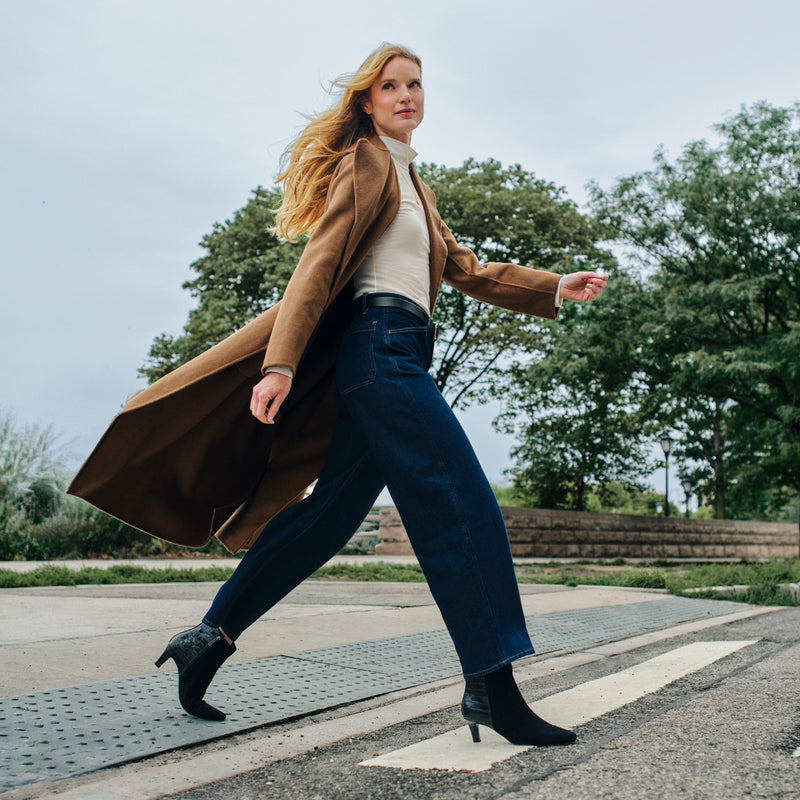 A woman walking outdoors on a paved path wearing a long brown coat, white top, dark blue wide-leg jeans, and black ankle boots.