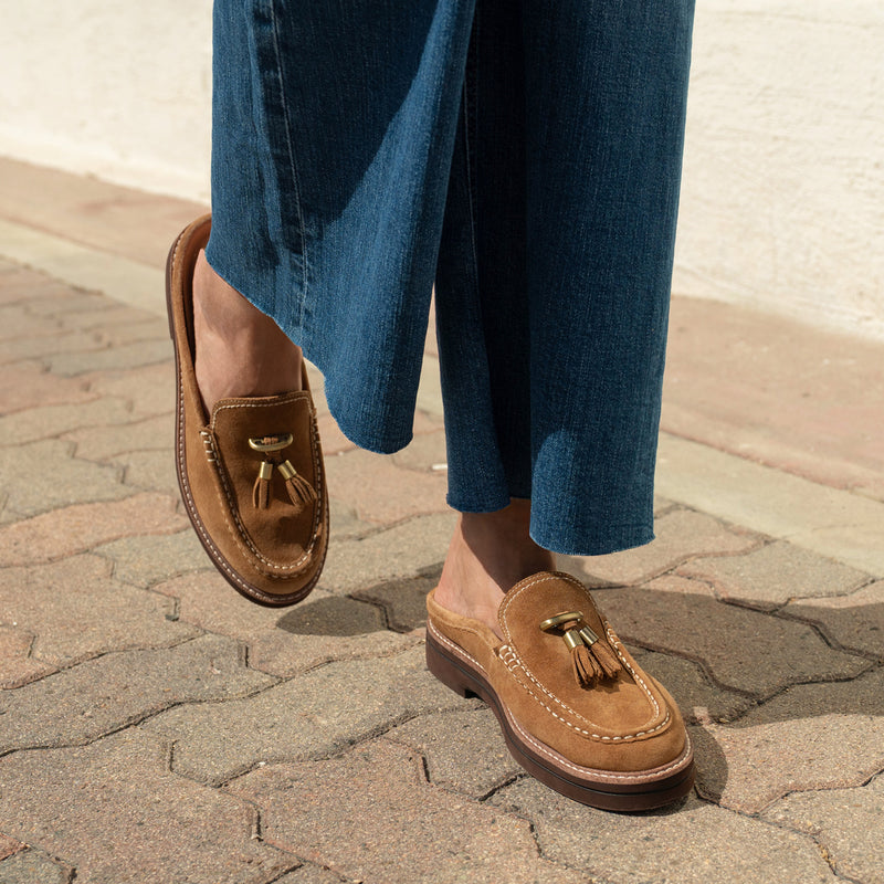 A person wearing tan suede tassel loafer mules with blue wide-leg jeans, standing on a paved outdoor walkway.