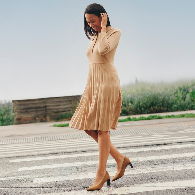 A woman wearing a beige pleated long-sleeve midi dress and matching beige heels walks across a zebra crossing outdoors.