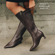 A person wearing dark brown knee-high leather boots with a pointed toe and low block heel, standing against a weathered gray concrete wall. White text on the right reads 'STRETCH PANEL FOR EASY FIT' with a dotted line pointing to the side zipper area.