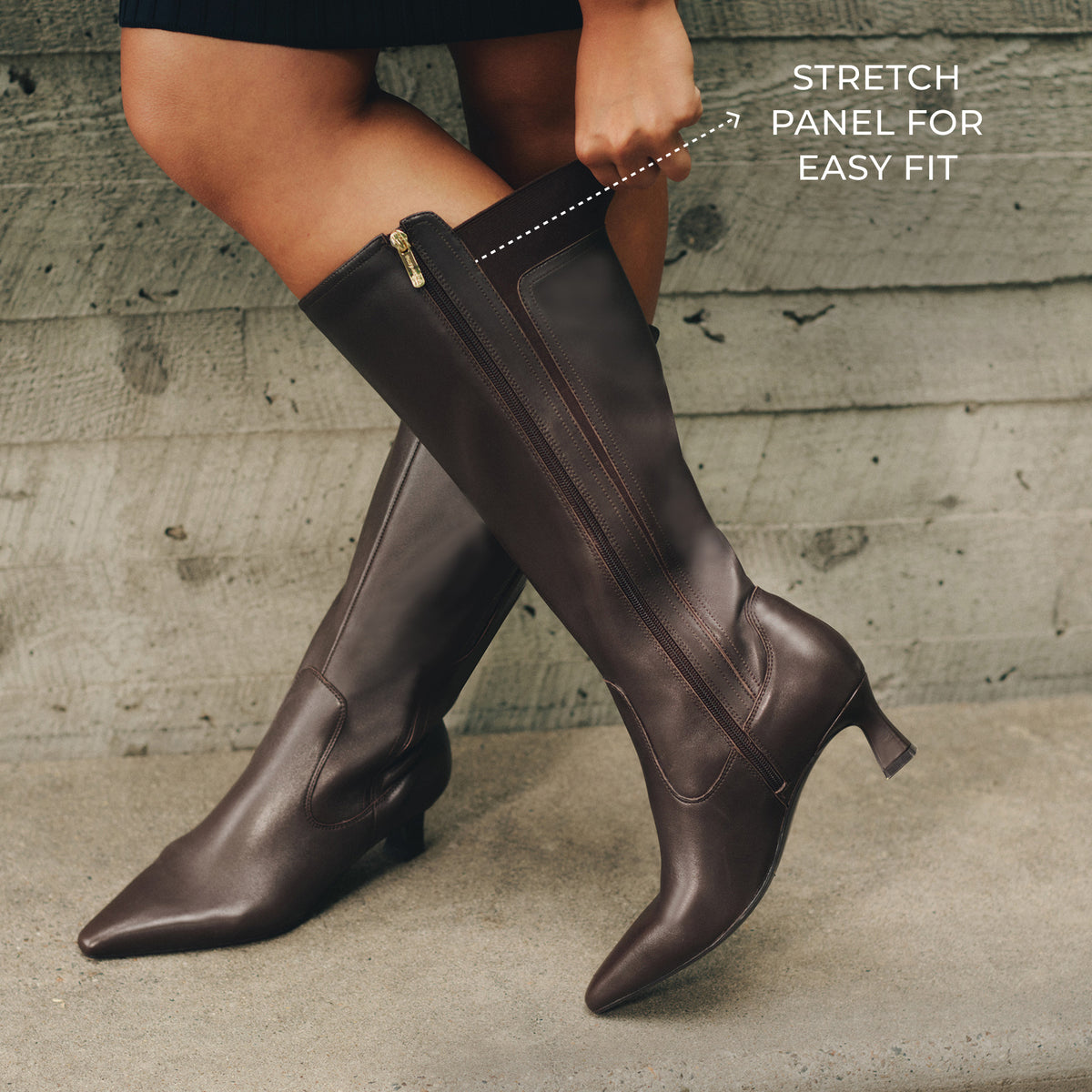 A person wearing dark brown knee-high leather boots with a pointed toe and low block heel, standing against a weathered gray concrete wall. White text on the right reads 'STRETCH PANEL FOR EASY FIT' with a dotted line pointing to the side zipper area.