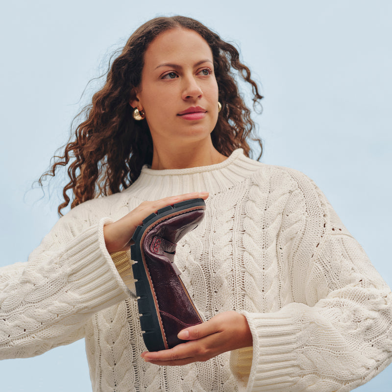 A woman with curly brown hair wearing a cream cable-knit sweater holds up a dark brown leather bootie to show its flexible sole. She is photographed against a solid light blue background.