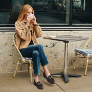 A woman sits on a cafe chair holding a white coffee cup, wearing a tan jacket, blue jeans, and brown loafers with horsebit details. She is seated next to a small metal cafe table on a concrete patio.
